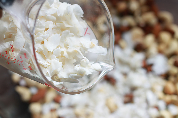 Mixing glass of dried coconut being poured into bowl filled with nuts.