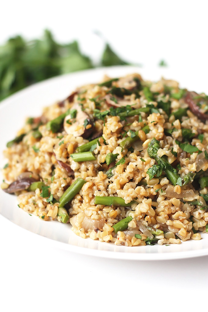 White plate filled with Freekeh with mushrooms and asparagus with green herbs in the background.