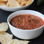 Bowl of salsa with tortilla chips around bowl and a bowl of chips in the background.