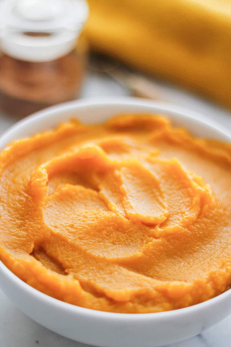 Bowl of smooth homemade pumpkin puree with a jar of homemade pumpkin pie spice and a golden napkin blurred in the background, ready for fall baking.
