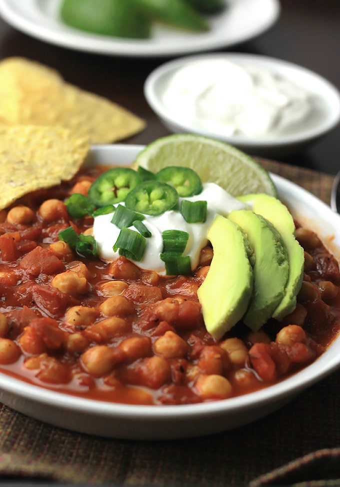 Bowl of Chickpea Chili topped with vegan sour cream, sliced avocado, jalapeno, scallions and a wedge of lime.