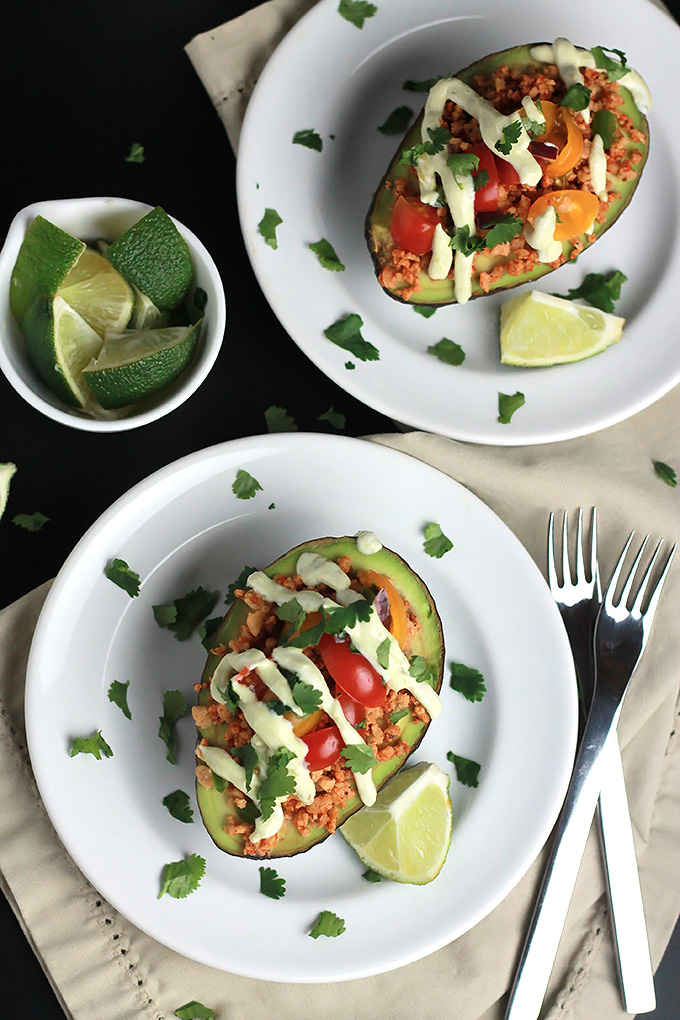 Overhead shot of sliced avocado stuffed with textured vegetable protein, topped with avocado cream, tomatoes and cilantro, with a wedge of lime.