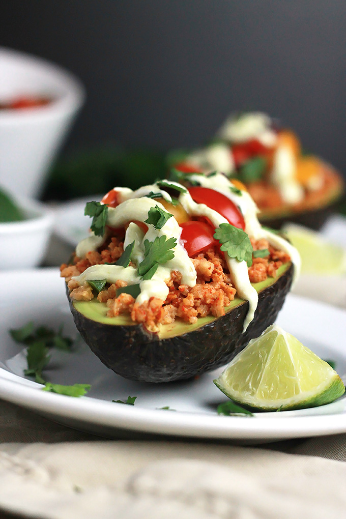 Close-up shot of sliced avocado stuffed with textured vegetable protein, topped with avocado cream, tomatoes and cilantro, with a wedge of lime.