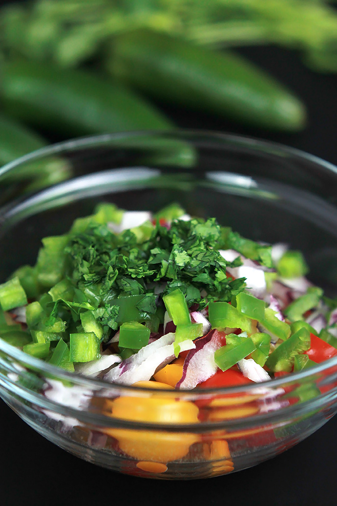 Glass bowl filled with salsa ingredients. Whole jalapenos and cilantro in background.