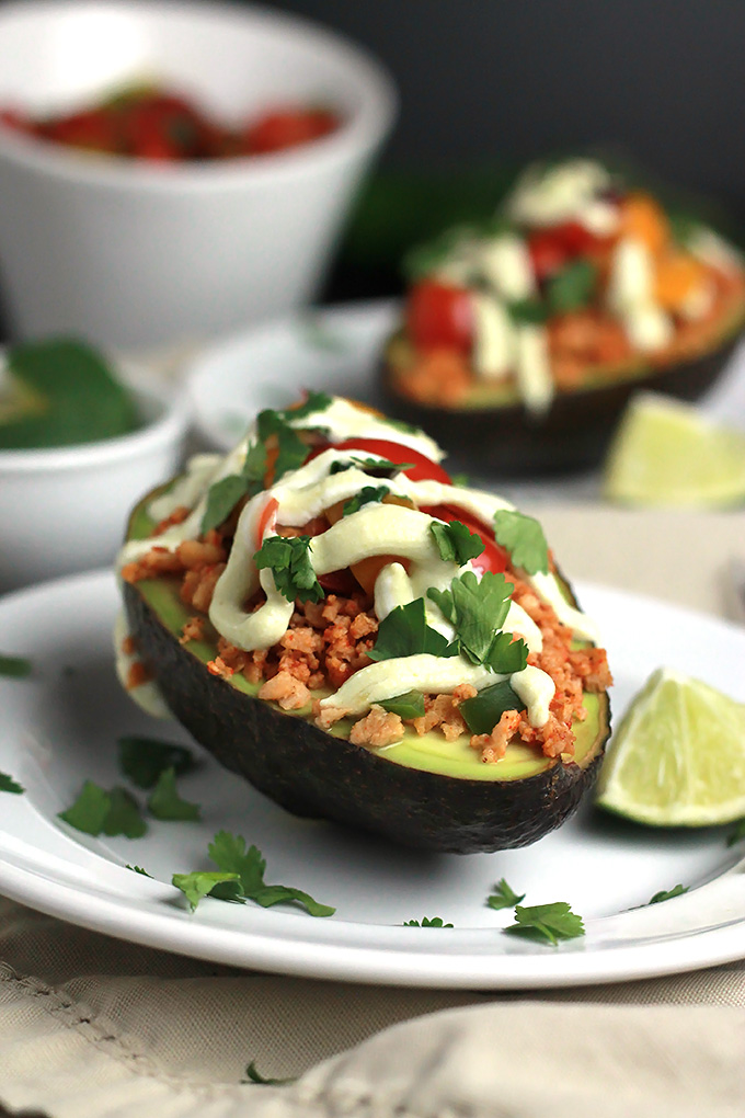 Sliced avocado stuffed with textured vegetable protein, topped with avocado cream, tomatoes and cilantro, with a wedge of lime.