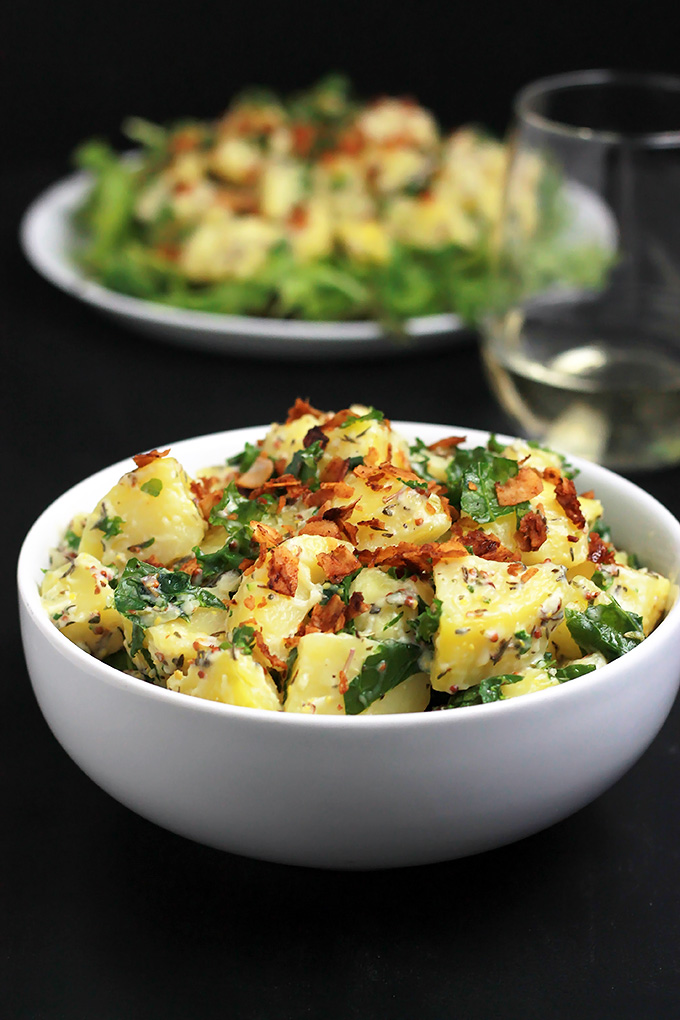 Vegan Yukon Kale Potato Salad in a bowl with the potato salad on a bed of arugula with coconut bacon on top in the background.