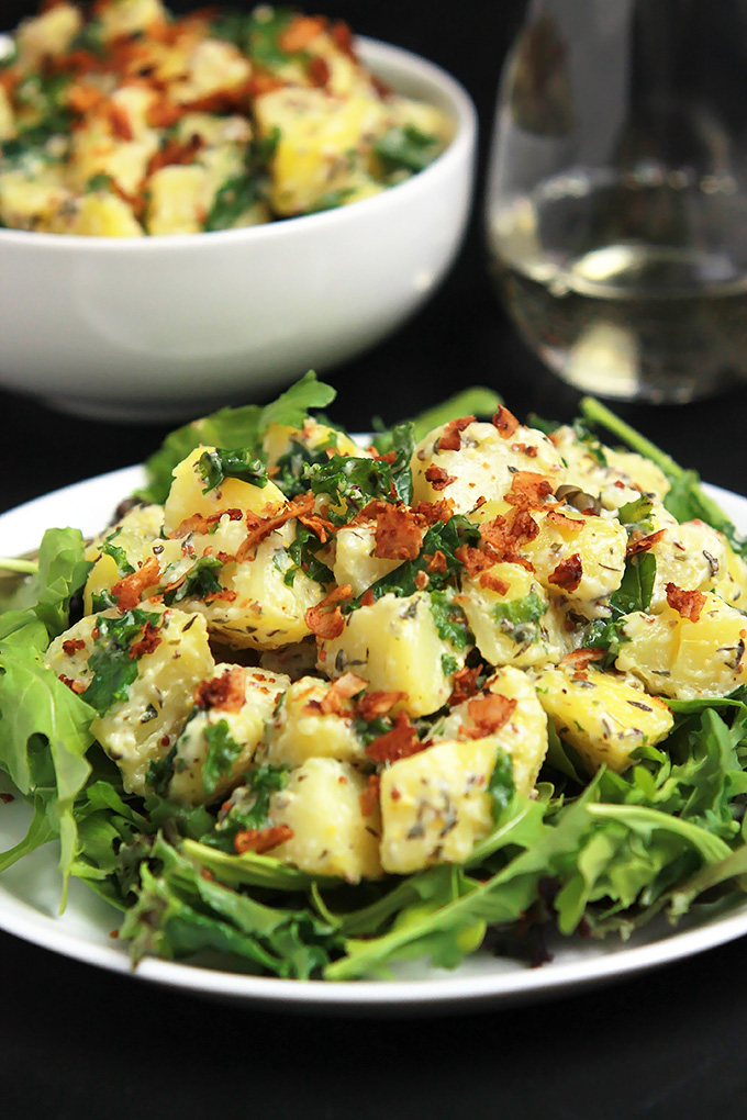 Vegan Yukon Kale Potato Salad on a bed of arugula with coconut bacon on top, bowl of potato salad is in the background.with a glass of white wine.