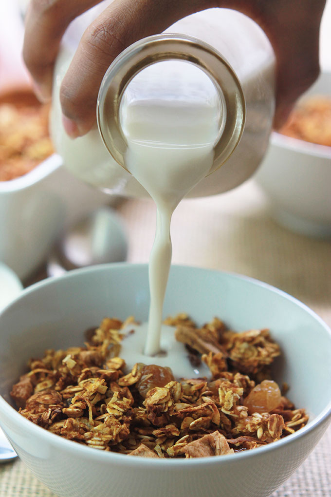 Pouring almond milk onto Pumpkin Apple Cinnamon Granola in a white bowl.