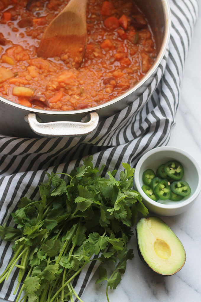 Pot of Spicy Lentil Sweet Potato Stew with a bunch of cilantro, sliced jalapenos and half of a sliced avocado beside pot of stew.