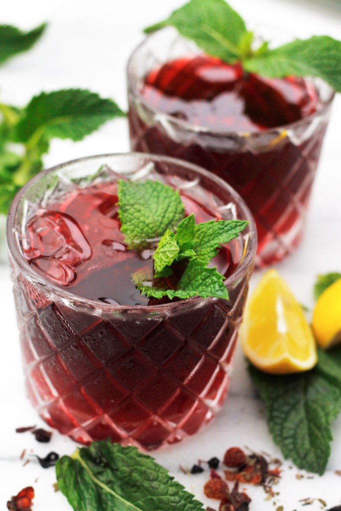 Two glasses of Sweet Vermouth Hibiscus Tea Cocktail garnished with fresh mint, with mint sprigs and sliced lemons beside the glass.