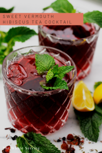 Two glasses filled with Hibiscus tea cocktail garnished with mint with lemon wedges on the side of glass.