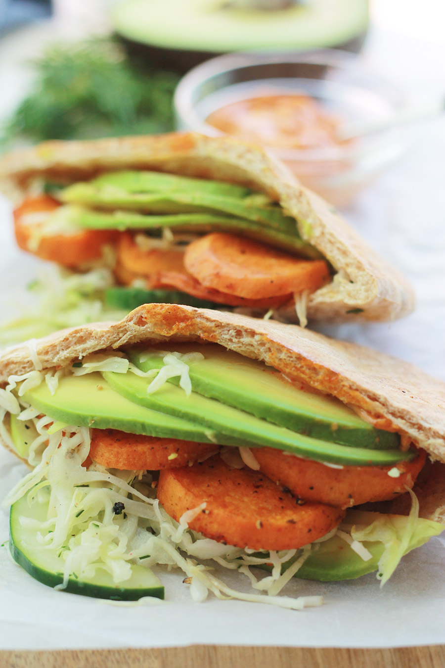 Pita pocket stuffed with spicy roasted sweet potato slices, cucumber slaw and avocado slices, with half of an avocado slice and sandwich spread in background.