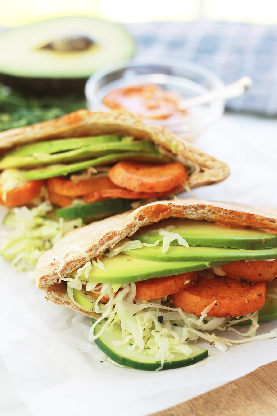 Pita pocket stuffed with spicy roasted sweet potato slices, cucumber slaw and avocado slices, with half of an avocado slice and sandwich spread in background.