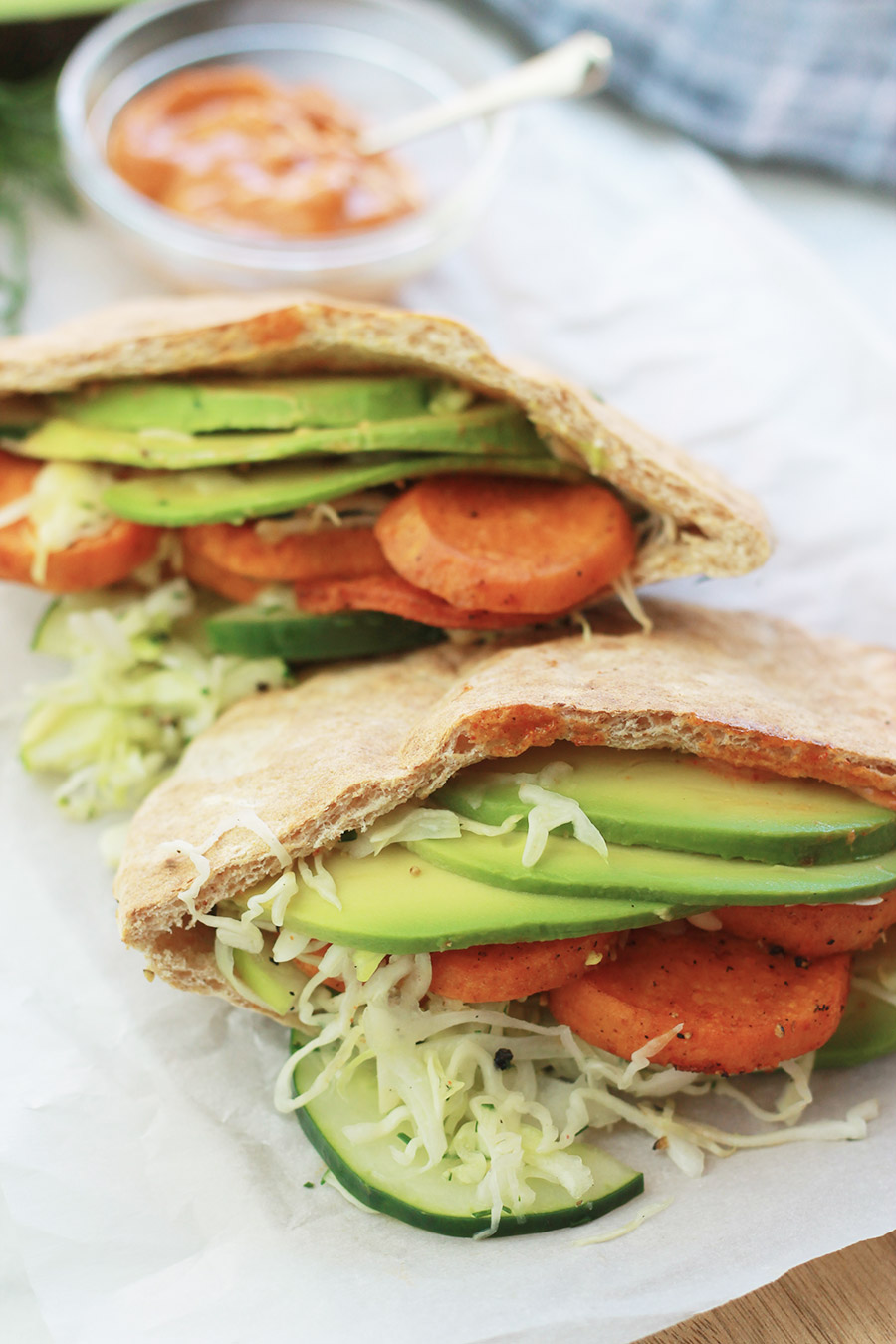 Close-up of pita pocket stuffed with spicy roasted sweet potato slices, cucumber slaw and avocado slices, with sandwich spread in background.