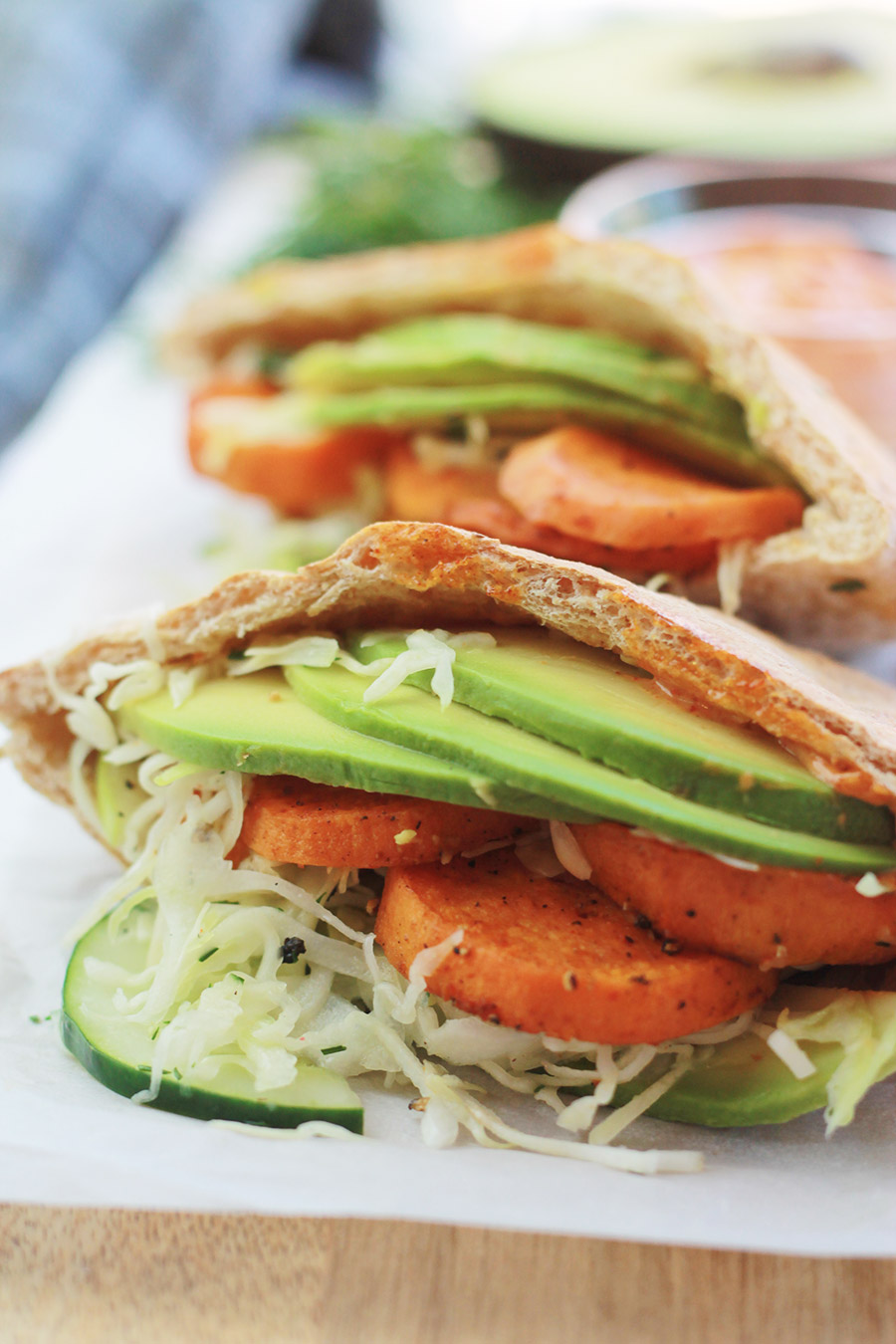 Close-up of pita pocket stuffed with spicy roasted sweet potato slices, cucumber slaw and avocado slices, with half of an avocado slice and sandwich spread in background.