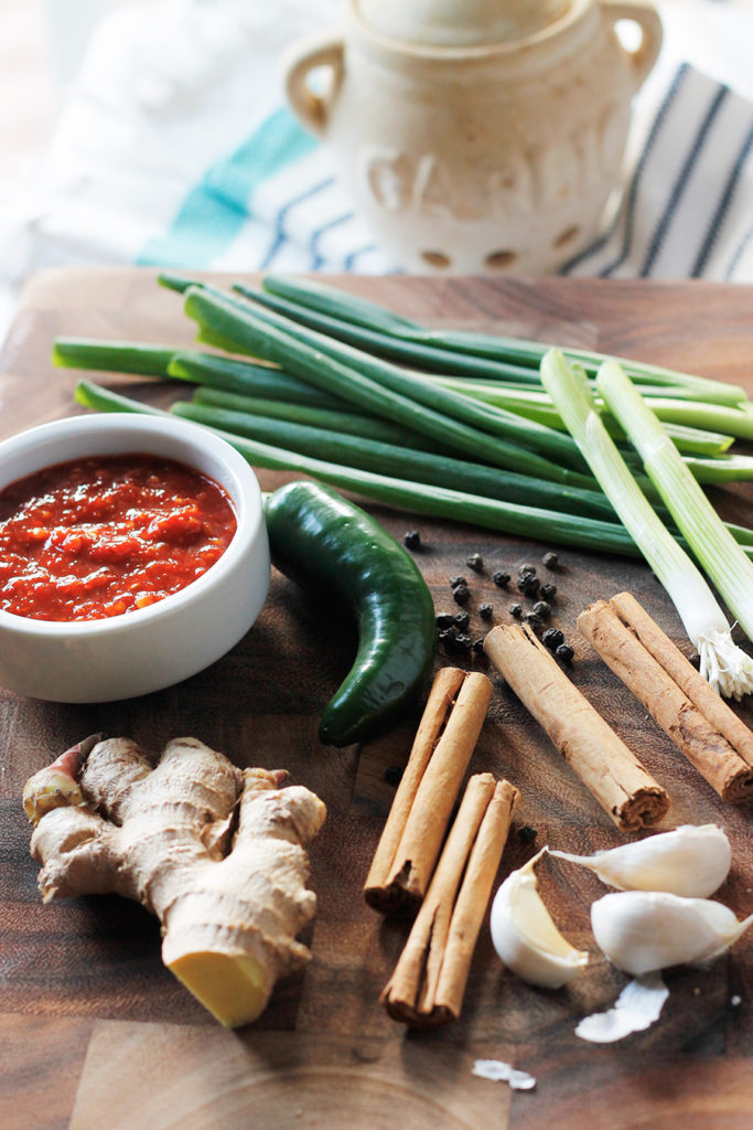 Ginger, cinnamon sticks, garlic, jalapeno, scallions and red chile sauce on wood cutting board.