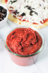 Bowl of Easy 5-Minute Pizza Sauce with a flatbread pizza and a bowl of black olives in background.