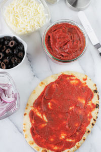 Overhead shot of Easy 5-Minute Pizza Sauce spread on flatbread with small individual bowls of pizza sauce, black olives, shredded cheese, and red onions to the left of the flatbread.