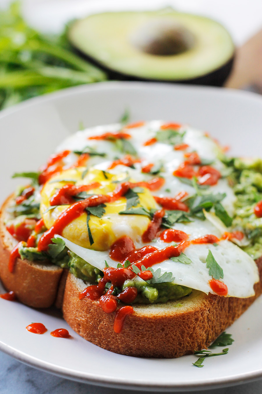 Avocado toast topped with fried egg sprinkled with torn fresh cilantro and drizzled with sriracha.