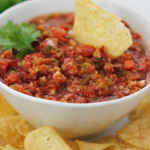 Bowl of salsa on a plate filled with tortilla chips and one tortilla chip dipped in salsa.