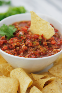Bowl of salsa on a plate filled with tortilla chips and one tortilla chip dipped in salsa.