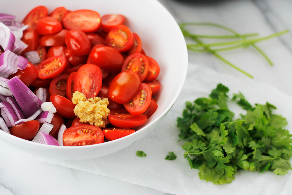 Sliced grape tomatoes, chopped red onions and minced garlic in a bowl with chopped cilantro beside the bowl.