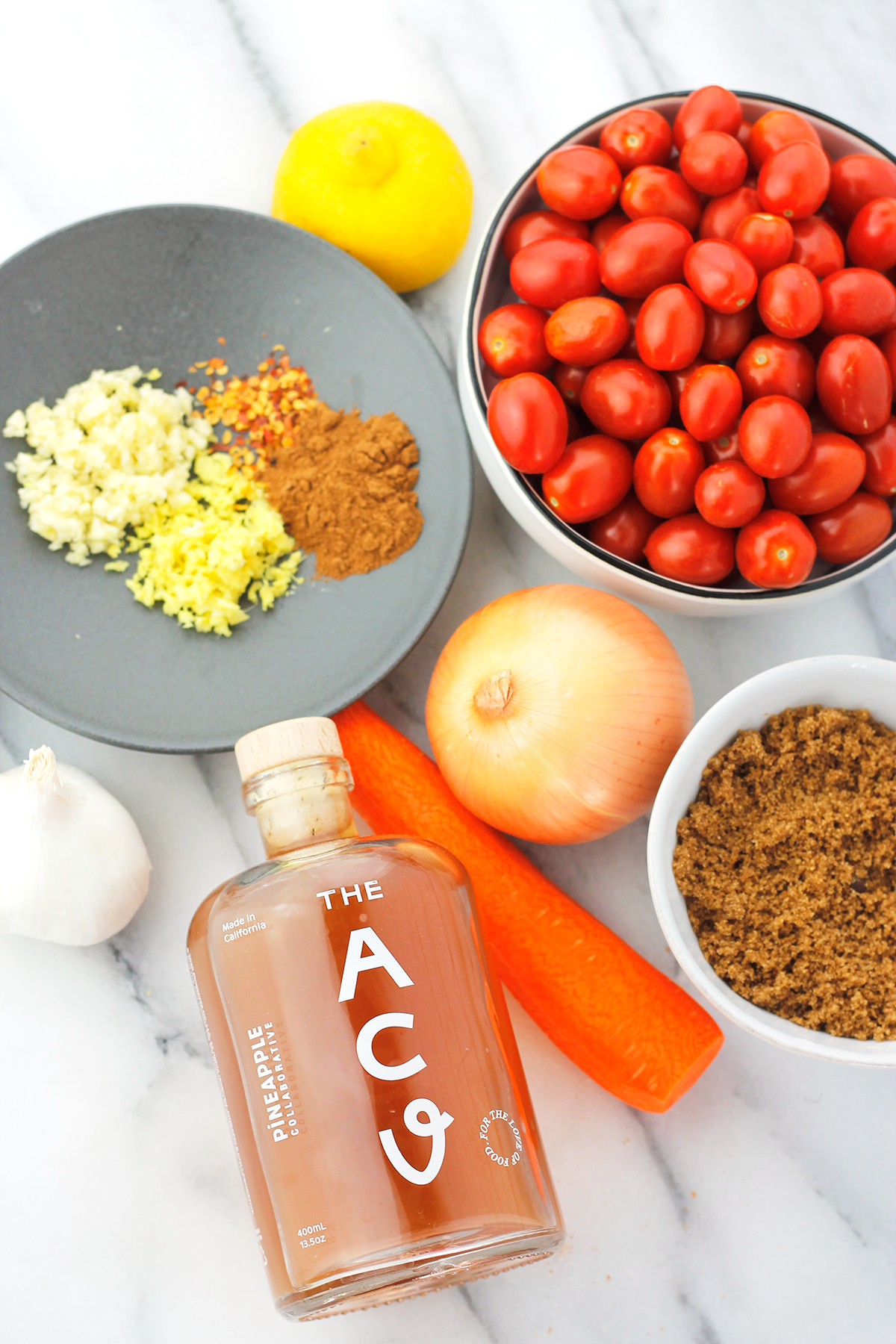 Ingredients needed to make chutney. Clockwise cherry tomatoes, sweet onion, brown sugar, carrot, jar of apple cider vineger, garlic, minced garlic, ginger, red pepper flakes and ground cinnamon on a saucer, and fresh lemon. 