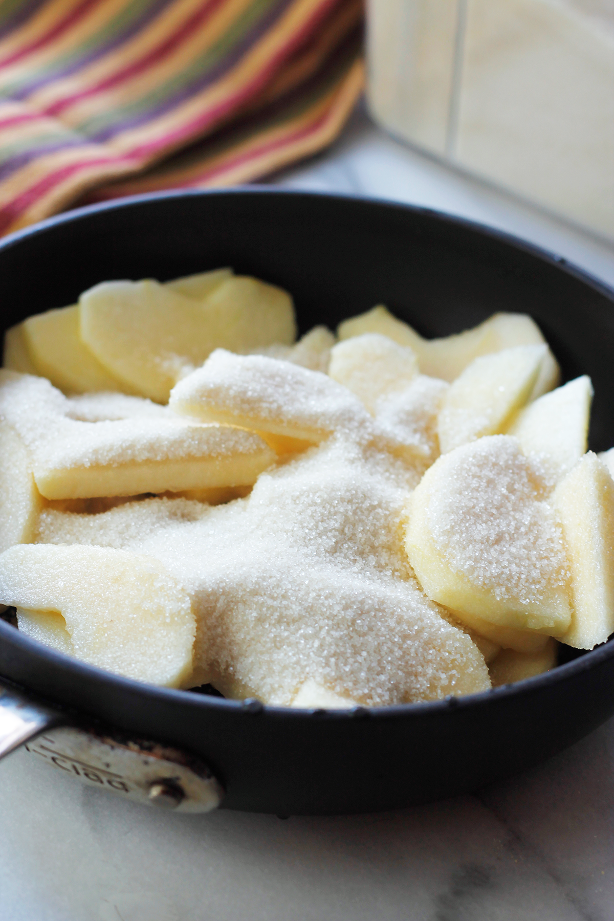 Pan filled with sliced apples covered in sugar and a striped kitchen towel in the background.
