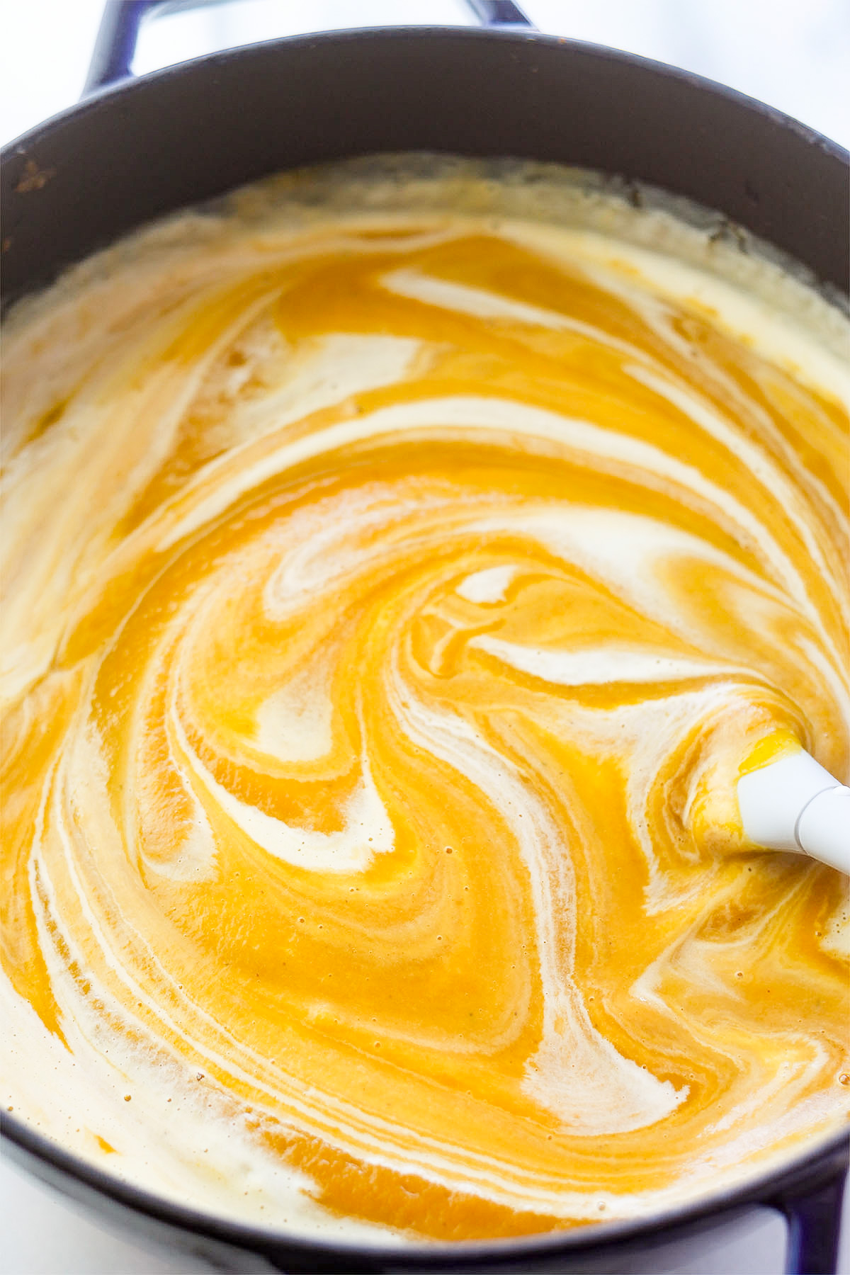 Overhead shot of butternut soup in a pot with swirls of heavy cream being mixed into the soup.