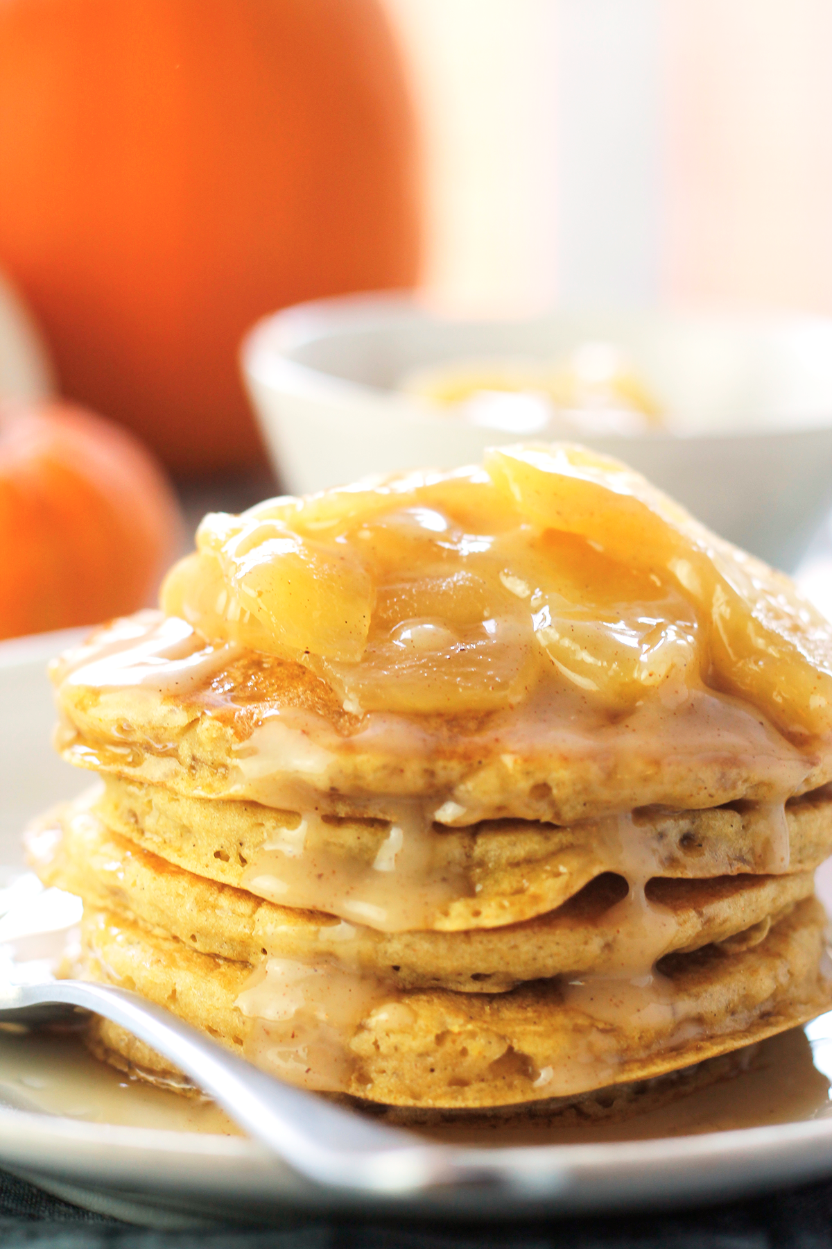 Stack of pumpkin pancakes on a plate with apples and pumpkin caramel sauce on top. A large orange pumpkin, medium white pumpkin and a small orange pumpkin in the background. A bowl of extra apple pumpkin sauce to the top right of the plate of pancakes.