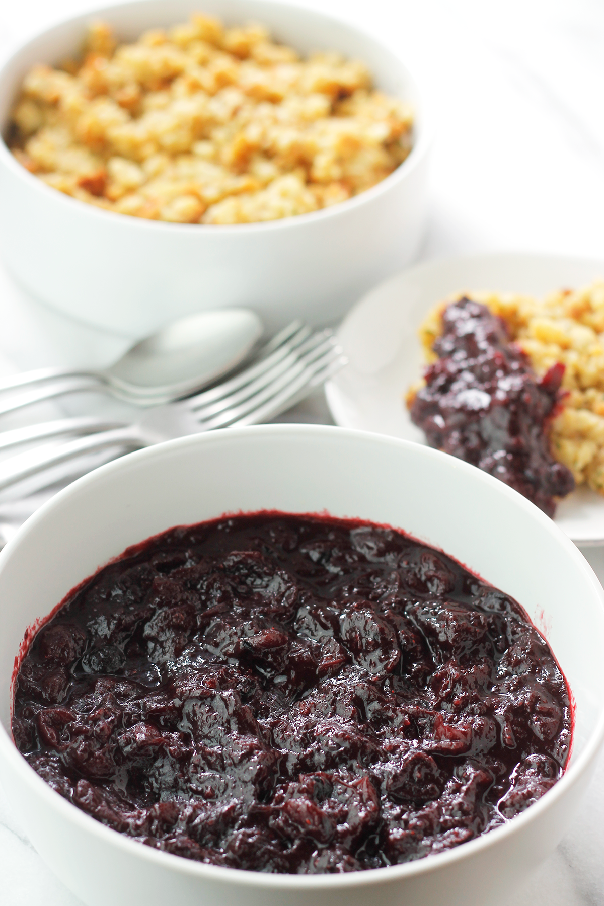 Bowl of cran-blueberry sauce with a bowl of stuffing and flatware in the background, and a small plate of stuffing with some cran-blueberry sauce  spooned over the stuffing.