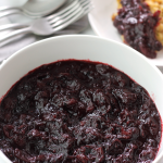 Bowl of cran-blueberry sauce with a bowl of stuffing and flatware in the background, and a small plate of stuffing with some cran-blueberry sauce spooned over the stuffing.