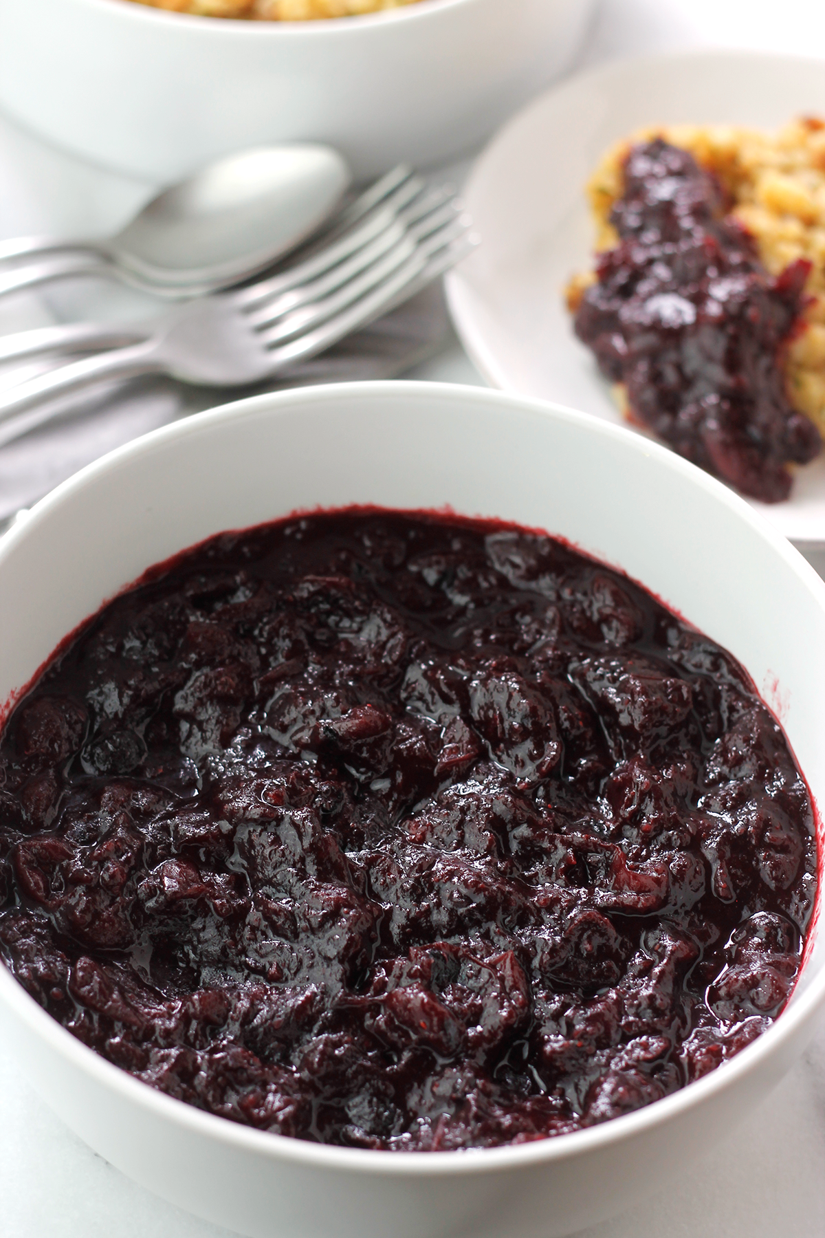 Bowl of cran-blueberry sauce with a bowl of stuffing and flatware in the background, and a small plate of stuffing with some cran-blueberry sauce spooned over the stuffing.