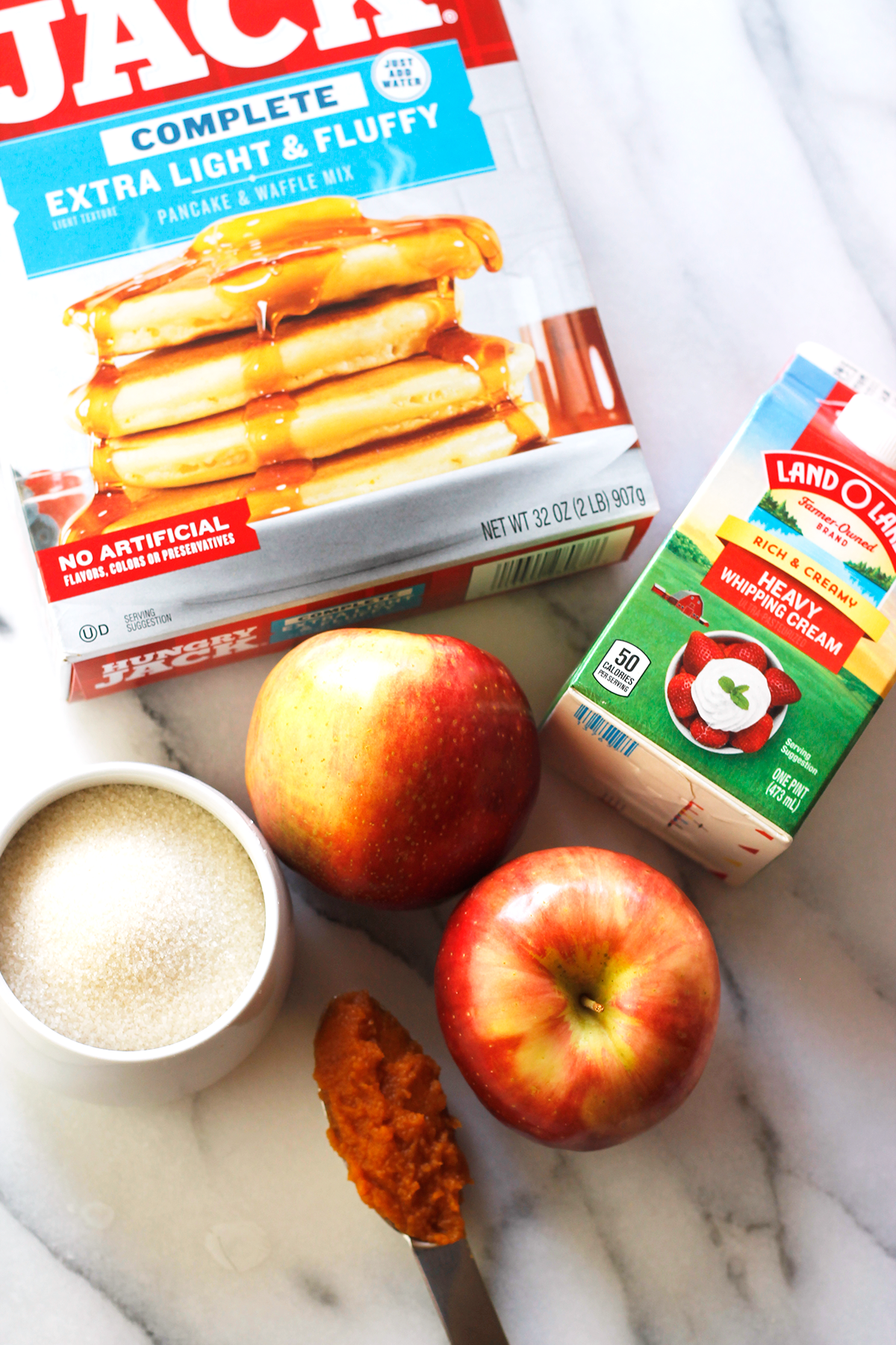 Overhead shot of the ingredients needed to make pumpkin pancakes with apples and pumpkin caramel sauce. Clockwise from the top, a box of complete pancake mix, carton of heavy whipping cream, two apples, tablespoon filled with pumpkin puree, and a bowl of sugar.