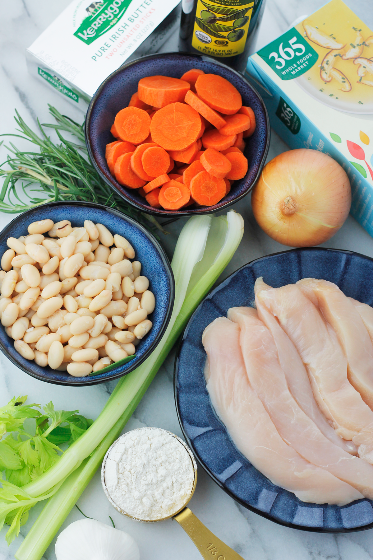 Overhead shot of ingredients for making creamy rosemary chicken soup. Clockwise butter, bottle of olive oil, box of chicken stock, unpeeled onion, to the left of the onion bowl of sliced carrots, to the right of onion plate of sliced raw chicken breast, measuring cup filled with flour, garlic, celery stalks, bowl of northern beans, and stems of rosemary.