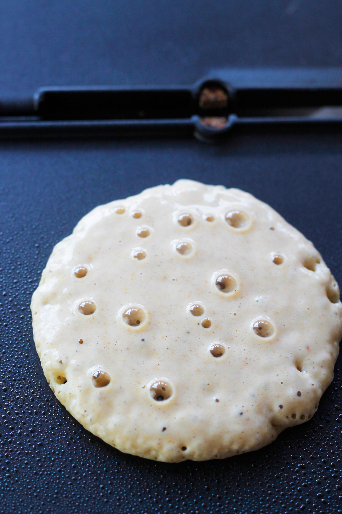 Pancake cooking on griddle with bubbles on top.