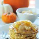 Stack of pumpkin pancakes on a plate with apples and pumpkin caramel sauce on top. A large orange pumpkin, medium white pumpkin and a small orange pumpkin in the background. A bowl of extra apple pumpkin sauce to the top right of the plate of pancakes.