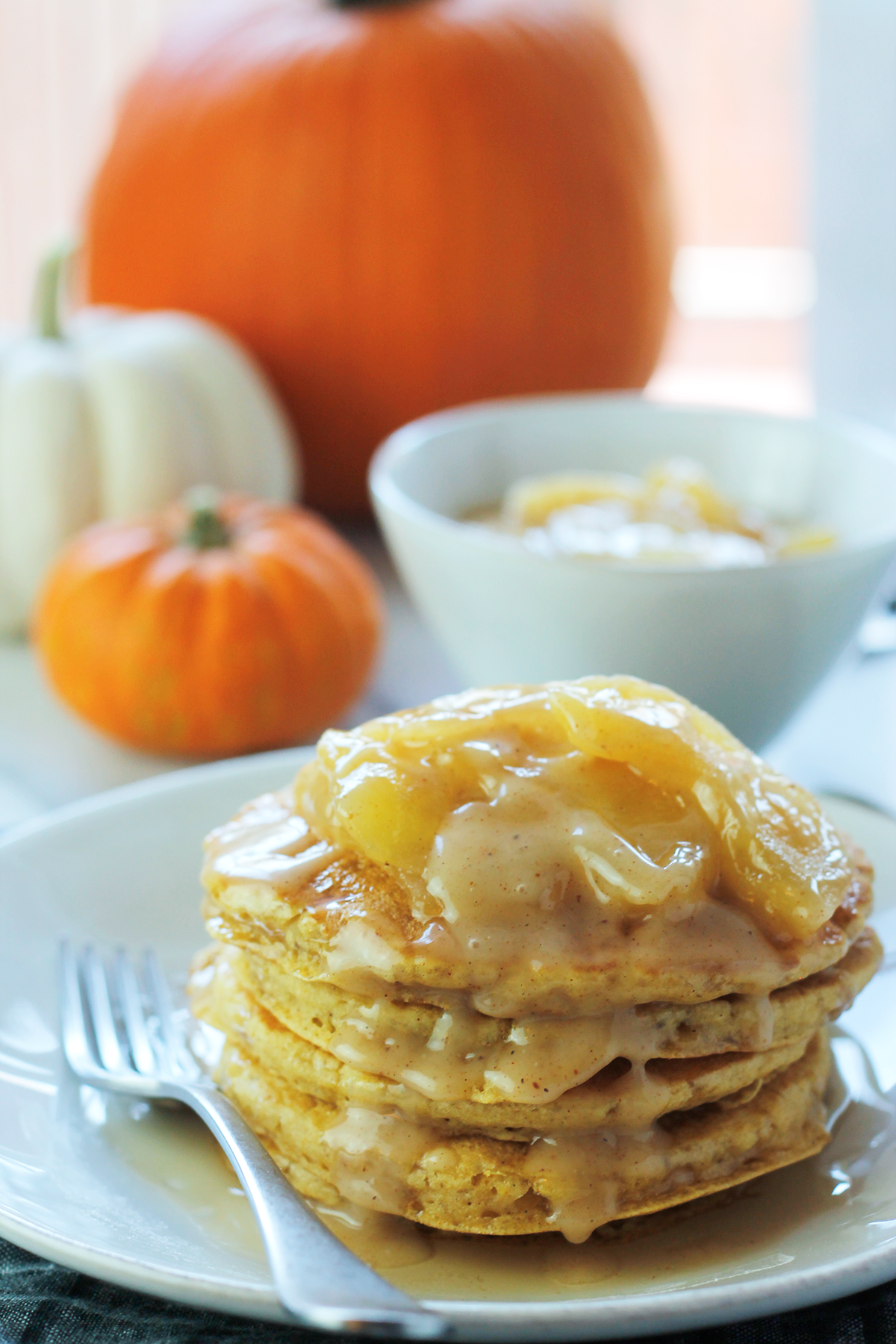 Stack of pumpkin pancakes on a plate with apples and pumpkin caramel sauce on top. A large orange pumpkin, medium white pumpkin and a small orange pumpkin in the background. A bowl of extra apple pumpkin sauce to the top right of the plate of pancakes. 