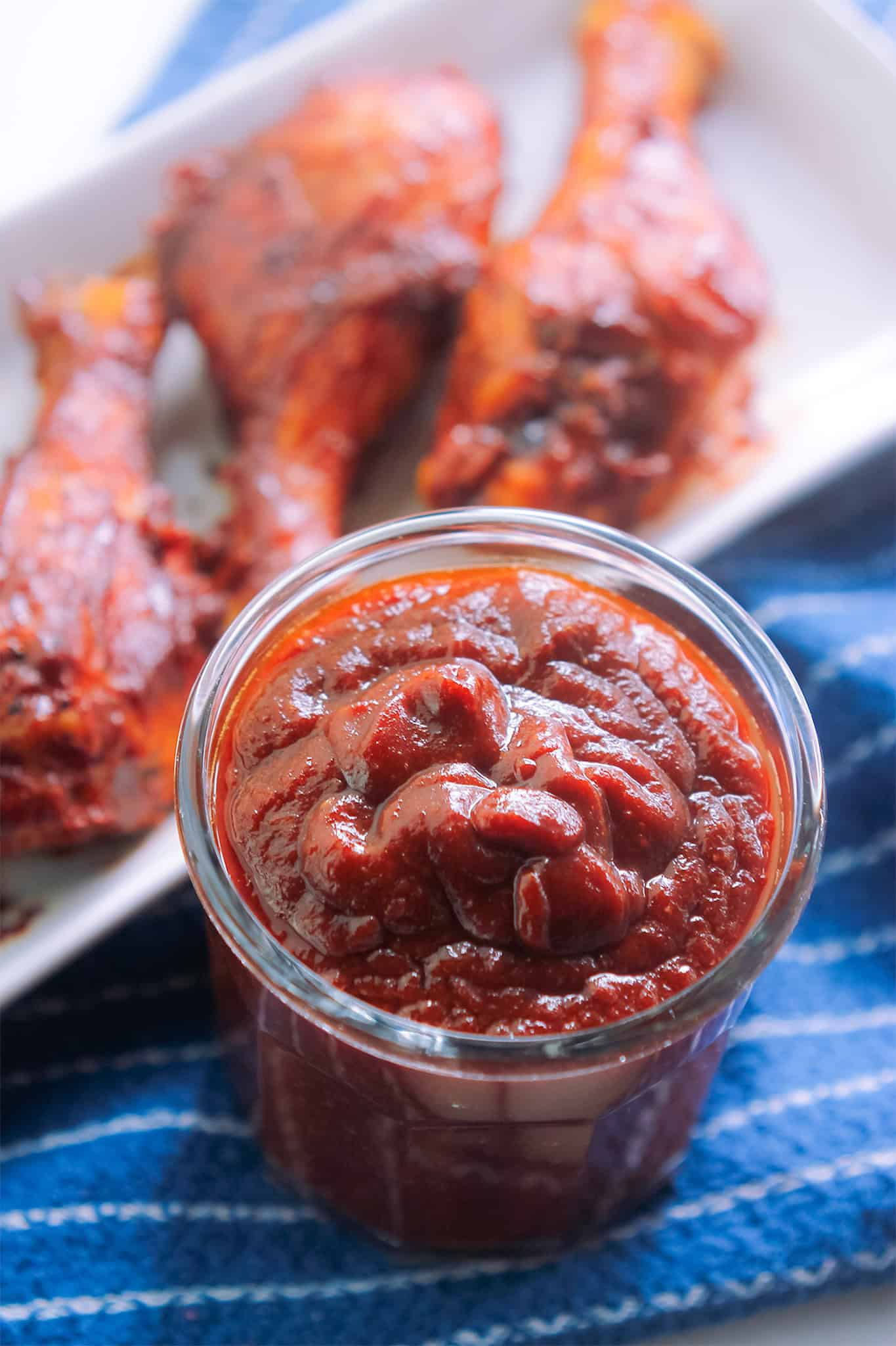 Glass container of prepared barbecue sauce with a platter of cooked barbecue drumsticks behind the barbecue sauce.