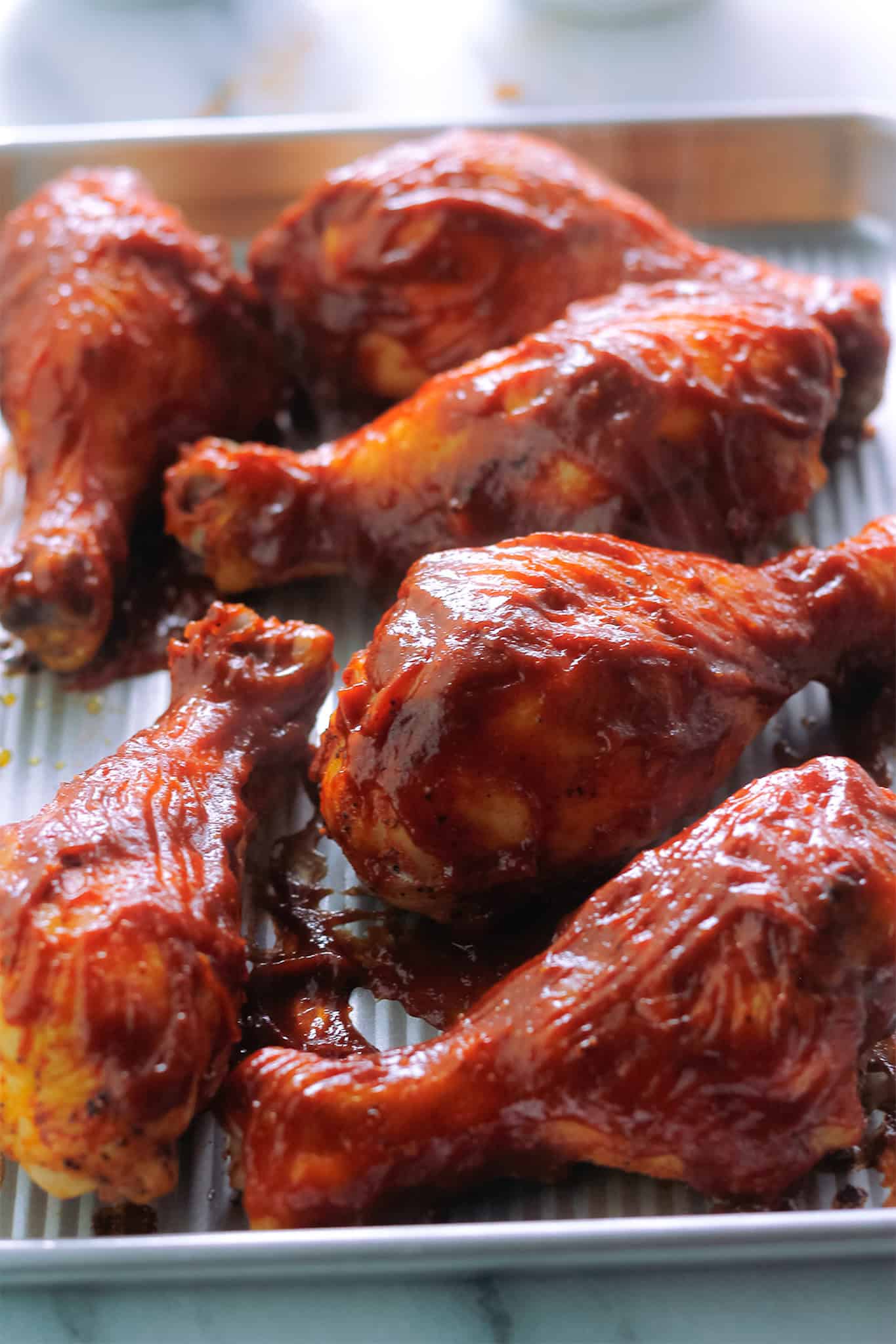 Partially cooked drumsticks smothered in barbecue sauce on sheet pan.