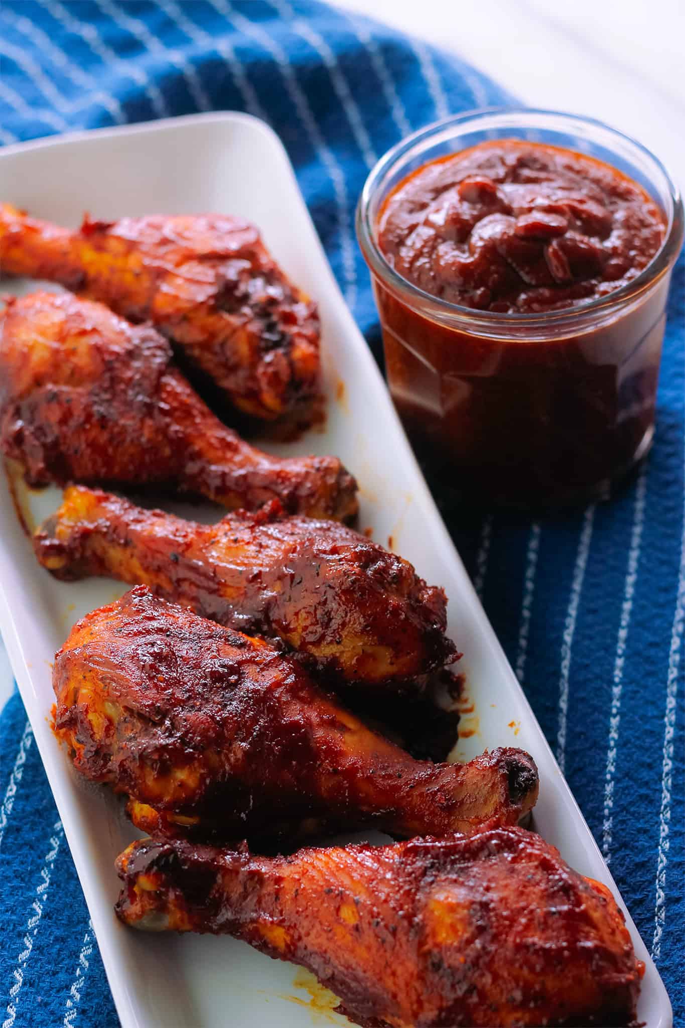 Platter of prepared drumsticks with a glass container of extra barbecue sauce on the right side of the platter.