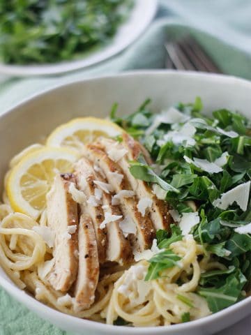 Overhead shot of lemon pasta with sliced grilled chicken and chopped arugula and a plate of more chopped arugula in the background.