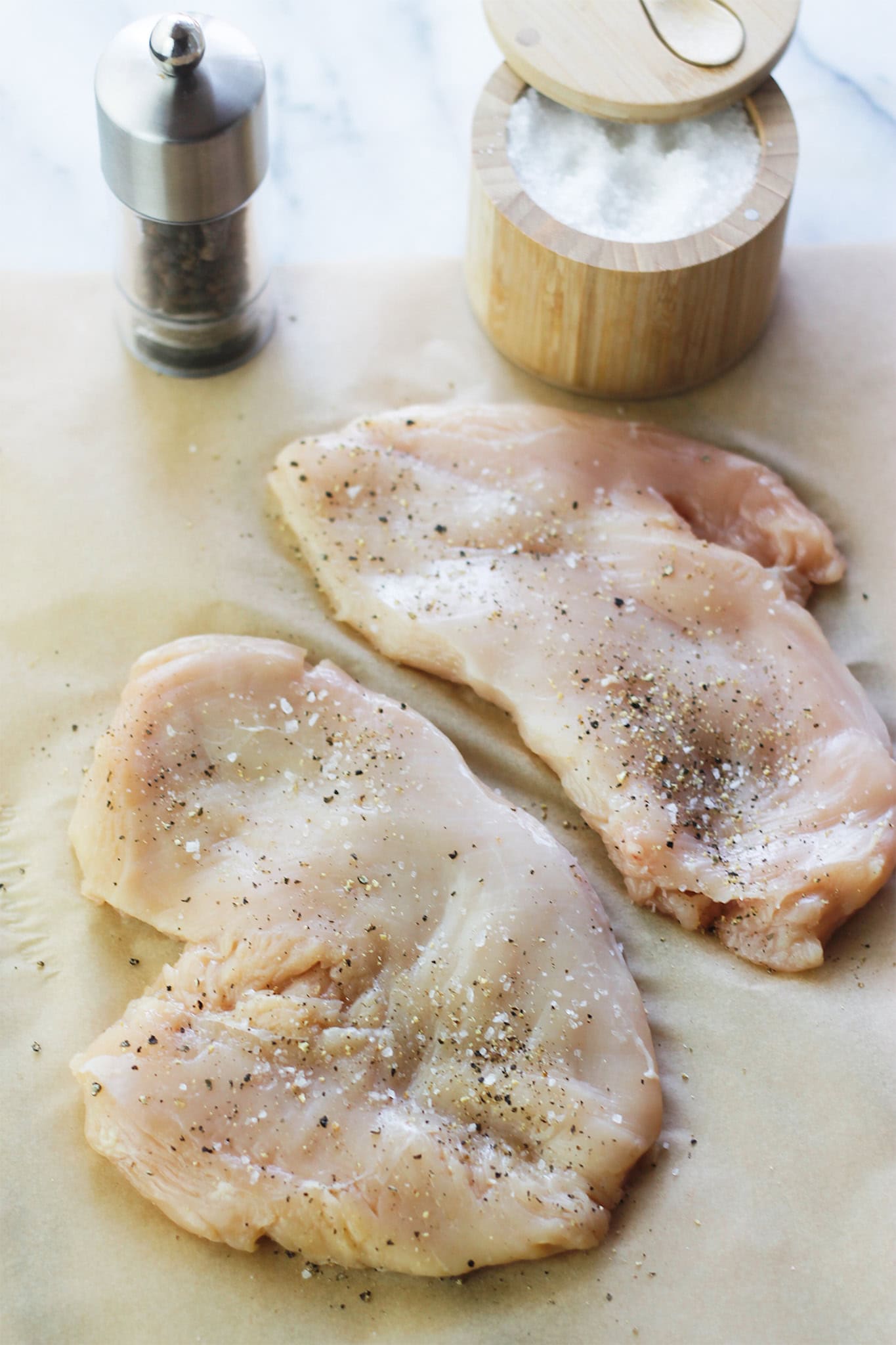 Two chicken breast being seasoned with salt and pepper.