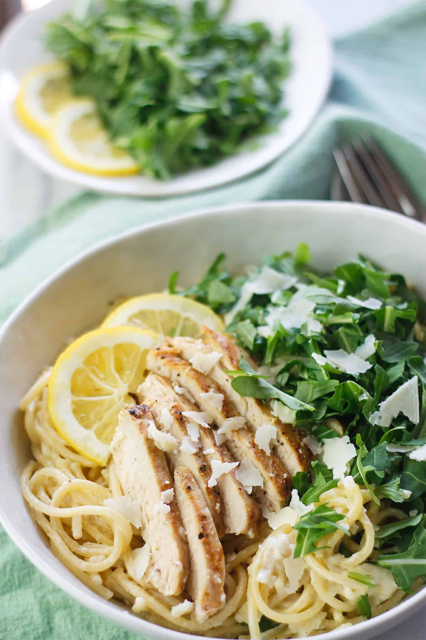 White bowl filled with pasta al limone topped with sliced grilled chicken and chopped arugula. There is a plate with sliced lemons and arugula in the background.