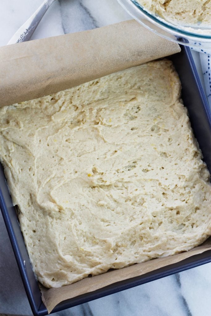 First layer of crumb cake batter spread on the bottom of a blue baking square pan lined with brown parchment paper.