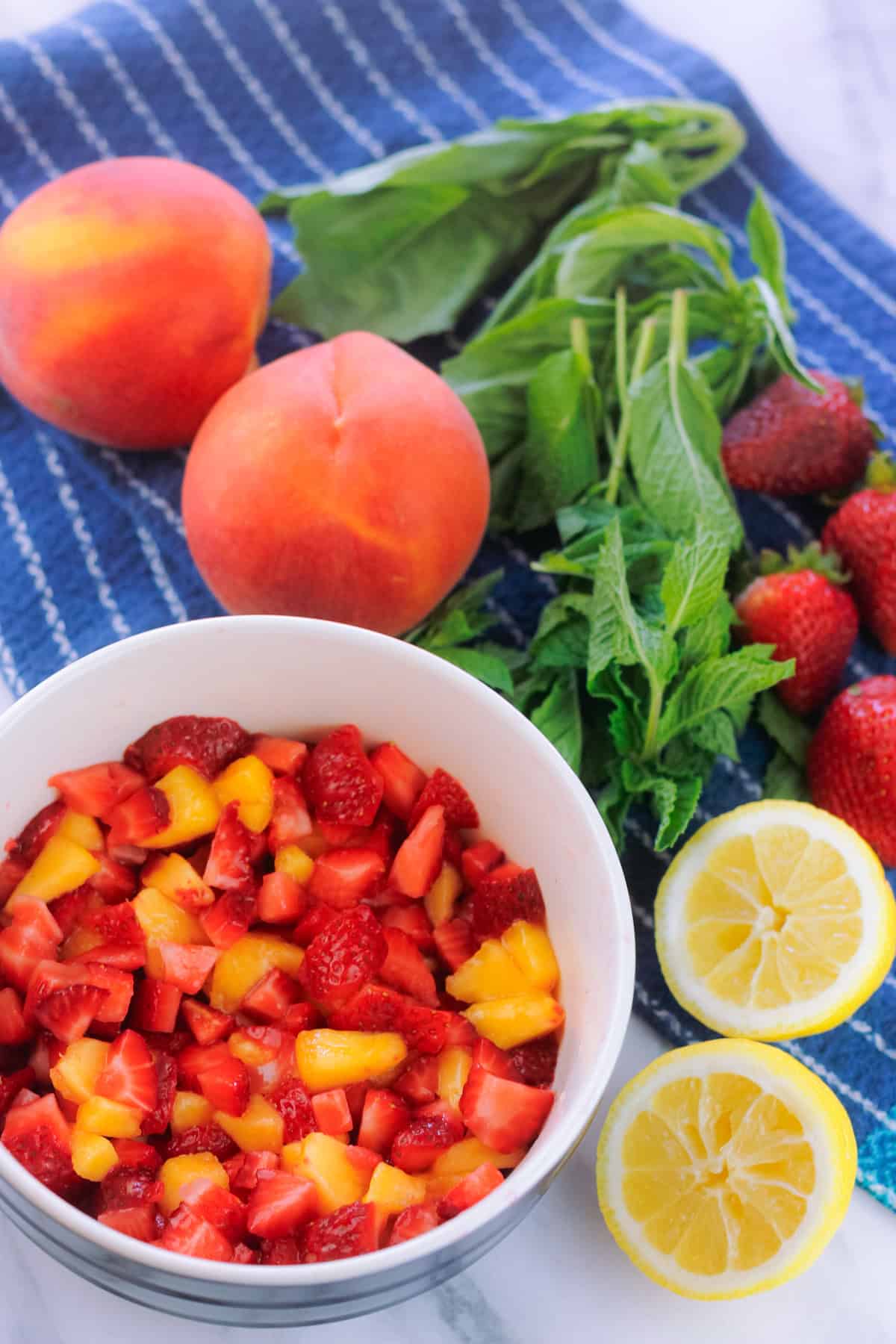 Bowl of diced strawberries and peaches with fresh cut lemon to the right of the bowl and fresh basil and mint and two peaches behind the bowl on a blue dish towel.