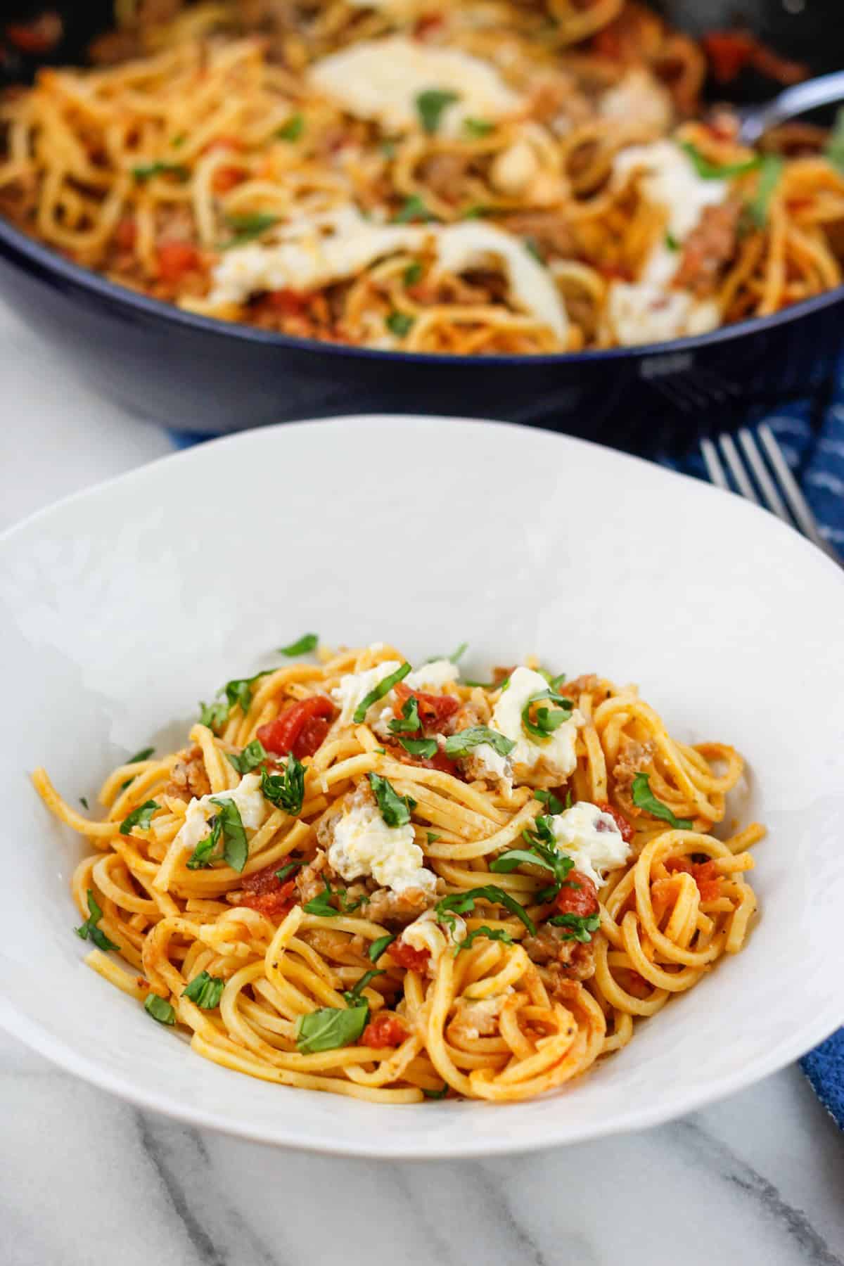 Bowl filled with sausage pasta with burrata and fresh basil on top with the skillet filled with the pasta in the background of the bowl of pasta.