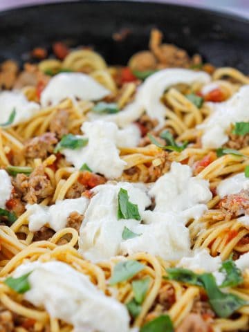 Close-up of sausage pasta with torn burrata scattered over the top and fresh torn basil leaves.