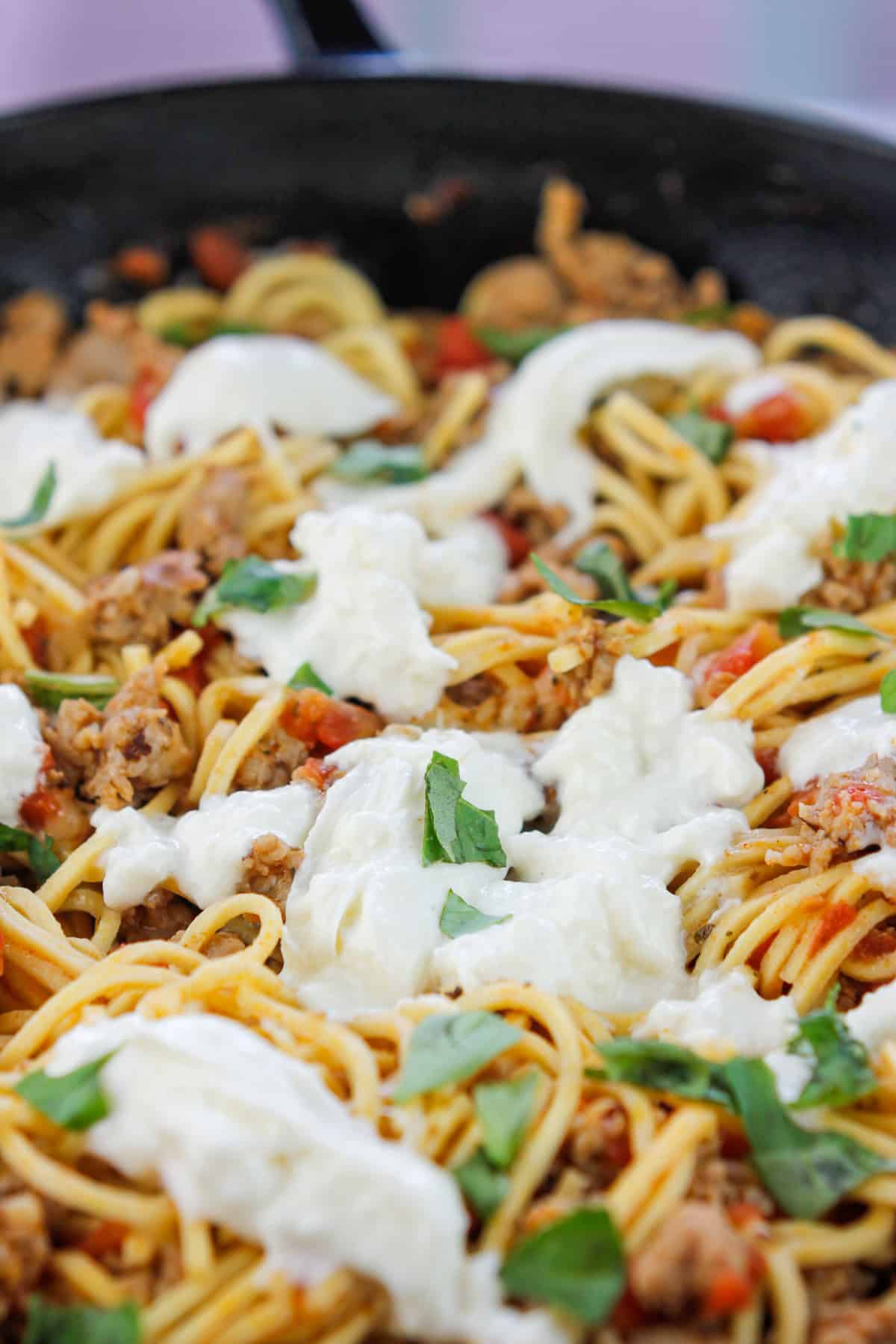 Close-up of sausage pasta with torn burrata scattered over the top and fresh torn basil leaves.