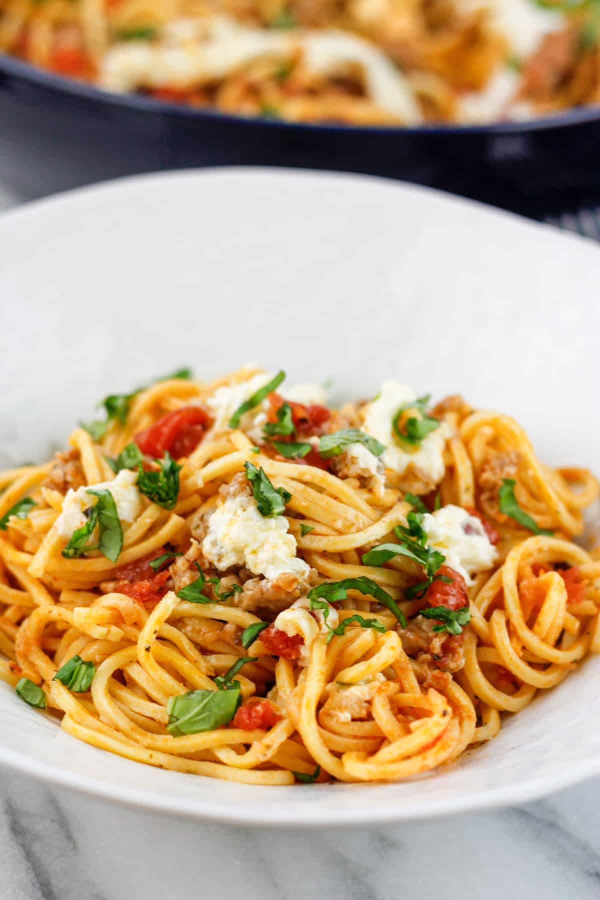 Close-up of bowl filled with sausage pasta with burrata and fresh basil on top with the skillet filled with the pasta in the background of the bowl of pasta.